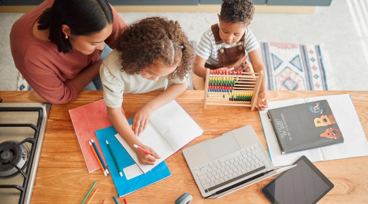 A Woman And Her Children Are Working At A Table With A Laptop.